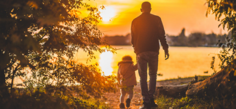 Photo of a man holding his daughter's hand as they walk towards the lake during sunset 